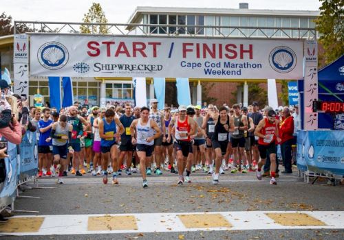 Runners gather at a Cape Cod Marathon start line under a banner reading &ldquo;START/FINISH&rdquo; in fall sunlight.