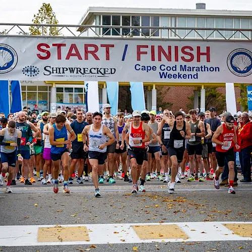 Runners gather at a Cape Cod Marathon start line under a banner reading &ldquo;START/FINISH&rdquo; in fall sunlight.