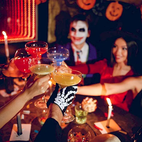 A Halloween party: guests with spooky makeup toast cocktails by candlelight around a decorated table.