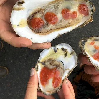 People hold open oysters with red roe, sharing bites and showing the shells on a street.
