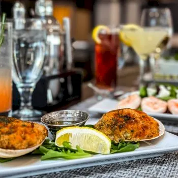 A restaurant table shows a plated fish cutlet/patty with a lemon wedge and greens, with drinks and other food in the background.