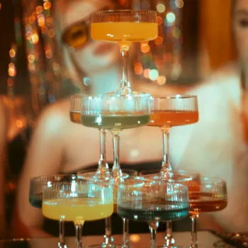 A bar setup with stacked cocktail glasses under warm bokeh lights, with two people blurred in the background.