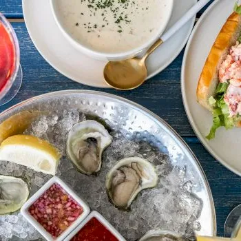 Oysters on ice with lemon, sauces and bread, plus a glass of red drink on a blue table.