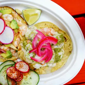 A plate of shrimp tacos with radish, cucumber, onion, and lime, topped with creamy green sauce on a bright red table.