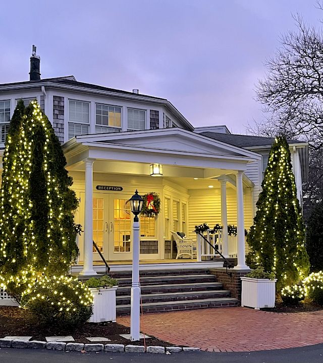 A hotel entrance at dusk, lit with glowing Christmas trees and warm lights, with a “RECEPTION” sign in the doorway.