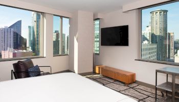 A modern hotel room with large windows showcasing a cityscape, a wall-mounted TV, armchair, small table, and a bed in the foreground.