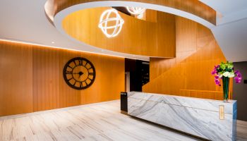 A modern lobby with a marble reception desk, wooden walls, a spiral staircase, a large wall clock, and ceiling lighting fixtures.