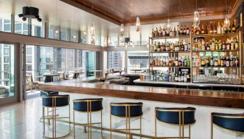 A modern bar with a wooden counter, blue cushioned bar stools, and a well-stocked liquor shelf, surrounded by large windows.
