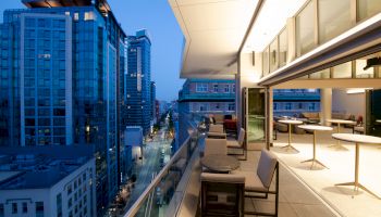 A modern cityscape view from a balcony at dusk, featuring high-rise buildings, an outdoor seating area, and an illuminated interior space.
