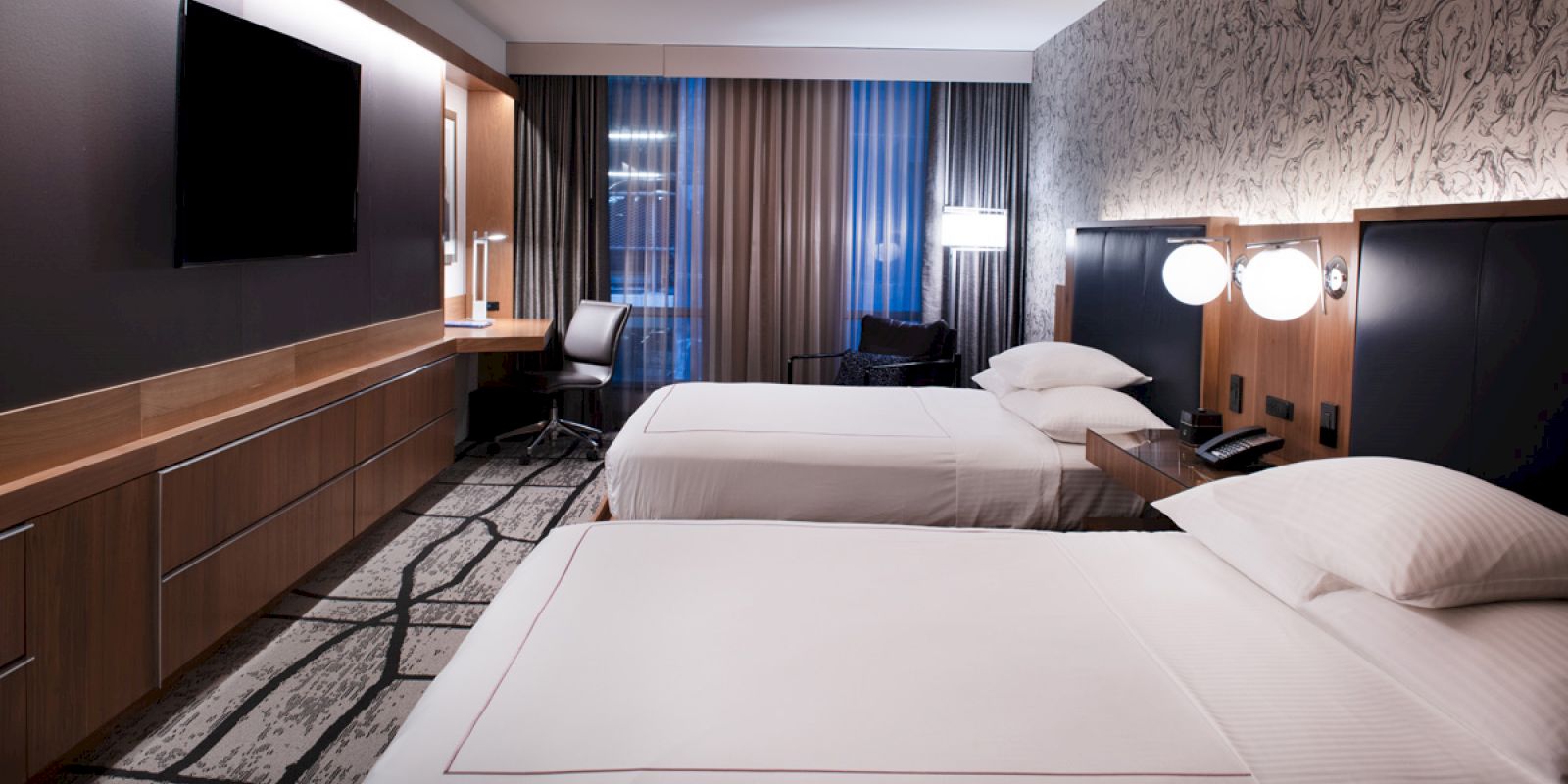 A modern hotel room features two neatly made beds, a wall-mounted TV, a work desk with a chair, stylish wall decor, and ambient lighting.