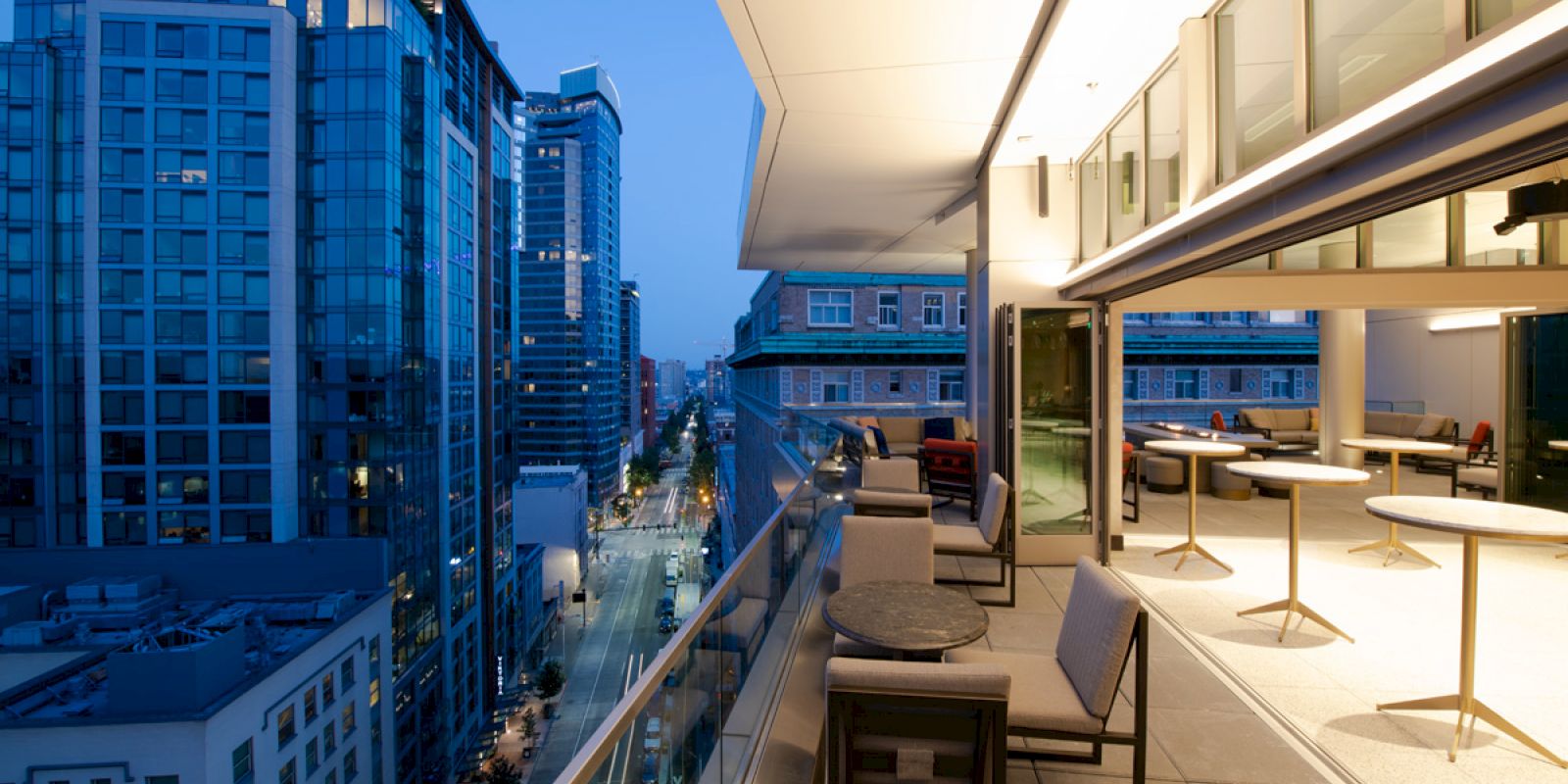 A cityscape at dusk featuring modern high-rise buildings. In the foreground, there's a spacious balcony with furniture and tables.