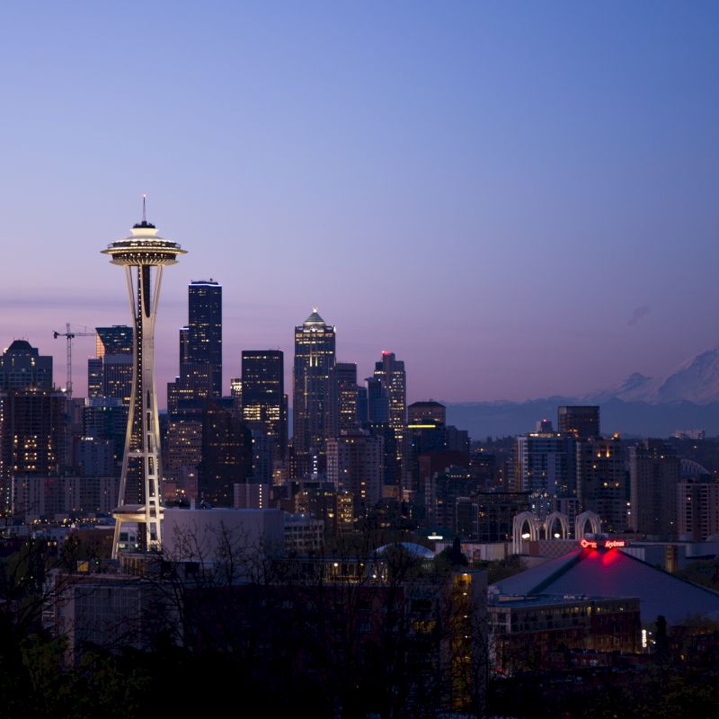 A twilight view of Seattle's skyline featuring the Space Needle, skyscrapers, and Mount Rainier in the distance, with a purple and pink sky.