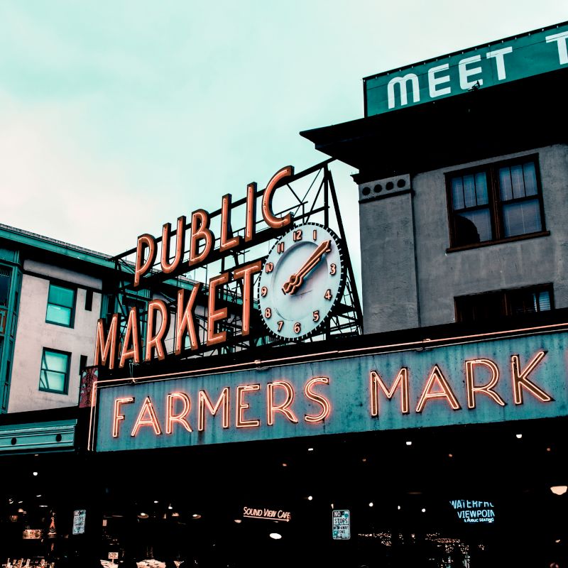 Image shows a neon sign for a "Public Market" and "Farmers Market" in front of buildings. A clock is prominent within the sign.