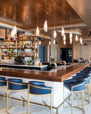 The image shows a modern bar with a wooden counter, blue chairs, hanging lights, and shelves full of bottles, creating a cozy atmosphere.