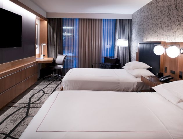 The image shows a modern hotel room with two double beds, a TV, desk, and chairs, featuring stylish decor and large windows with curtains.