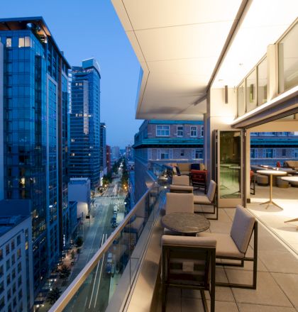 A modern high-rise apartment balcony at dusk, featuring outdoor seating and tables, with a view overlooking a busy city street and adjacent buildings.