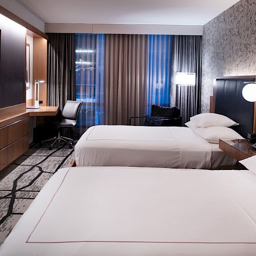 This image shows a modern hotel room with two neatly made beds, a wall-mounted TV, a desk with a chair, and stylish lighting fixtures.