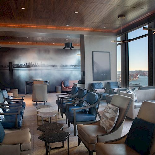 A modern lounge area with comfortable seating, large windows offering a city and waterfront view, and a mural on the back wall depicting a skyline.