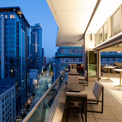 A cityscape at dusk, with modern high-rise buildings on the left and an open-air terrace with tables and chairs on the right side.