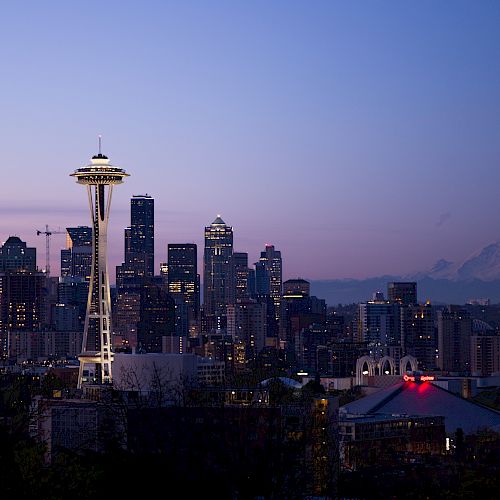 A cityscape of Seattle at dusk featuring the Space Needle, downtown skyscrapers, and Mount Rainier in the background, under a clear purple sky.