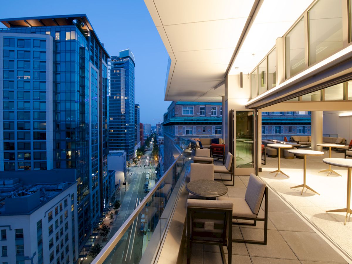 An urban balcony with modern furniture overlooks a city street lined with tall, illuminated buildings at dusk.