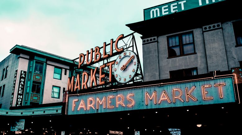 A neon sign reads "Public Market" and "Farmers Market" with a clock in between, against a backdrop of buildings including a hotel.