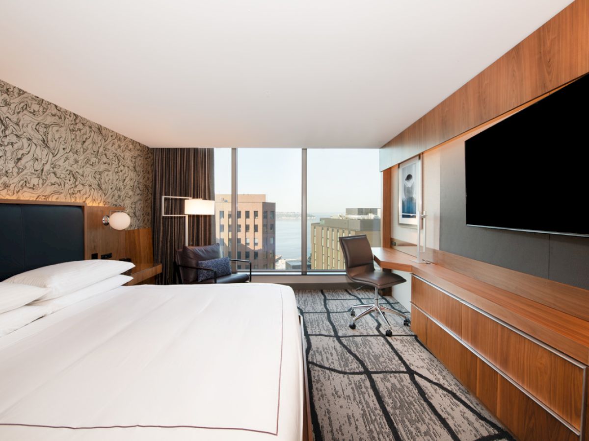 The image shows a modern hotel room with a large bed, a desk, a chair, a TV, and a window with a city view.