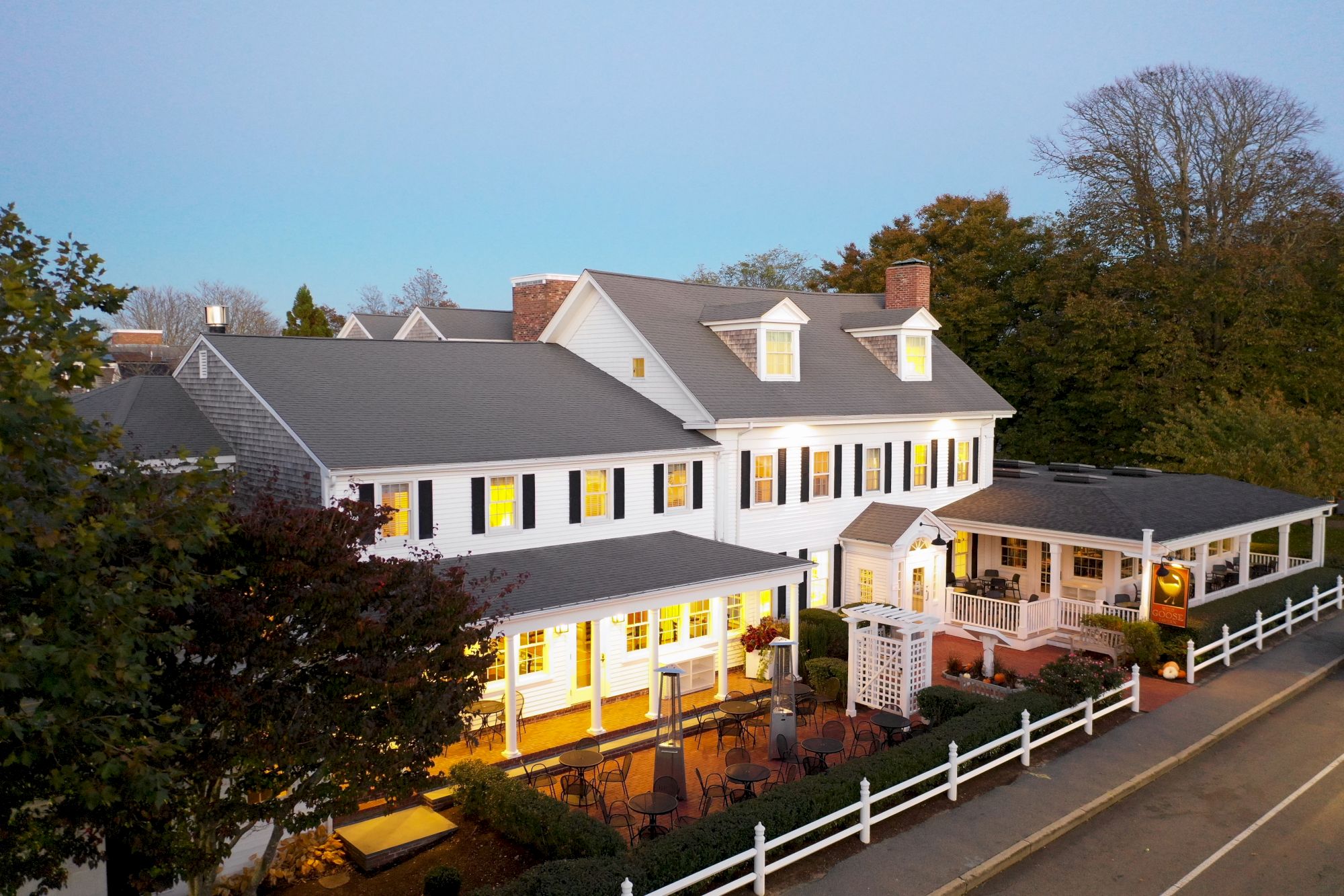 A two-story white building with a grey roof and illuminated windows, surrounded by trees, a white fence, and adjacent to a road.